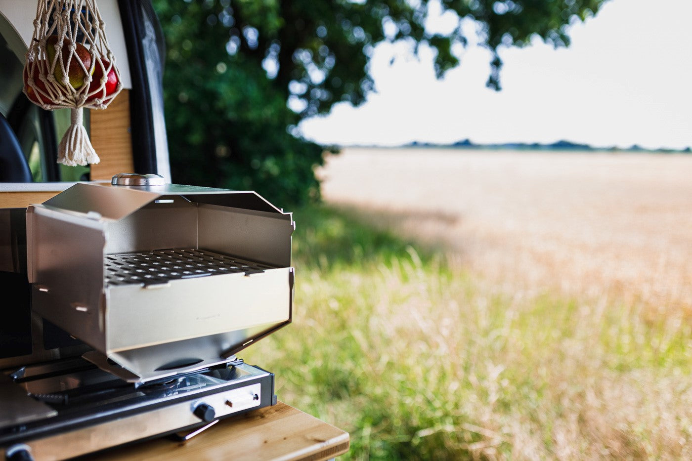 Ambientefoto:
Horst der multifunktionale Campingofen auf einem Gaskocher im Camper mit schönem Blick über ein Feld am Waldrand