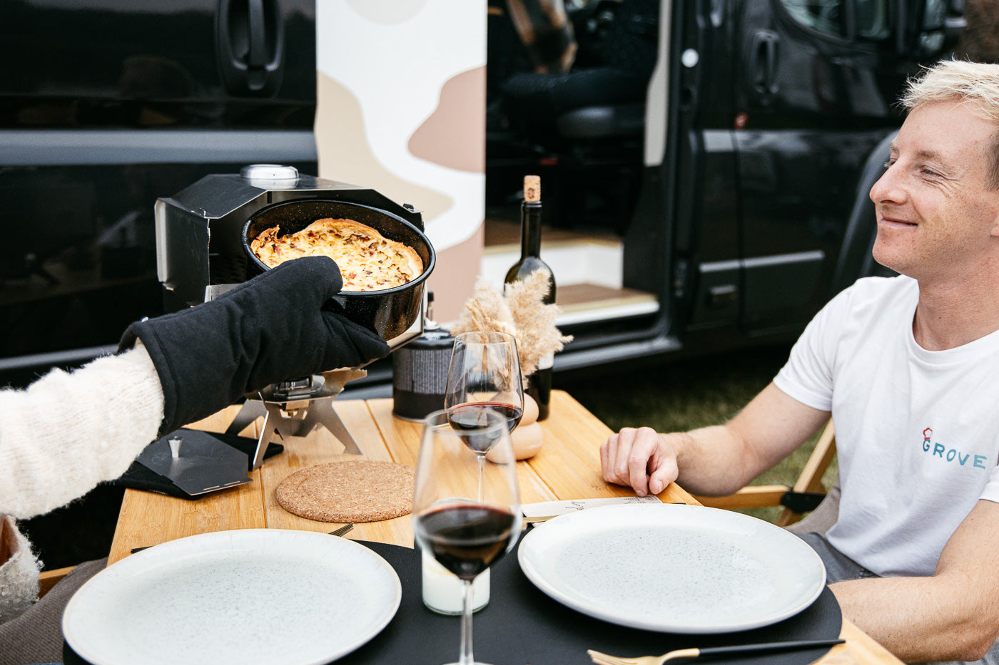 Ambientefoto:
Horst der multifunktionale Campingofen   als Campingofen auf Helga auf einem schön gedeckten Campingtisch vor einem Camper. Das fertige Essen wird grade aus dem Ofen genommen