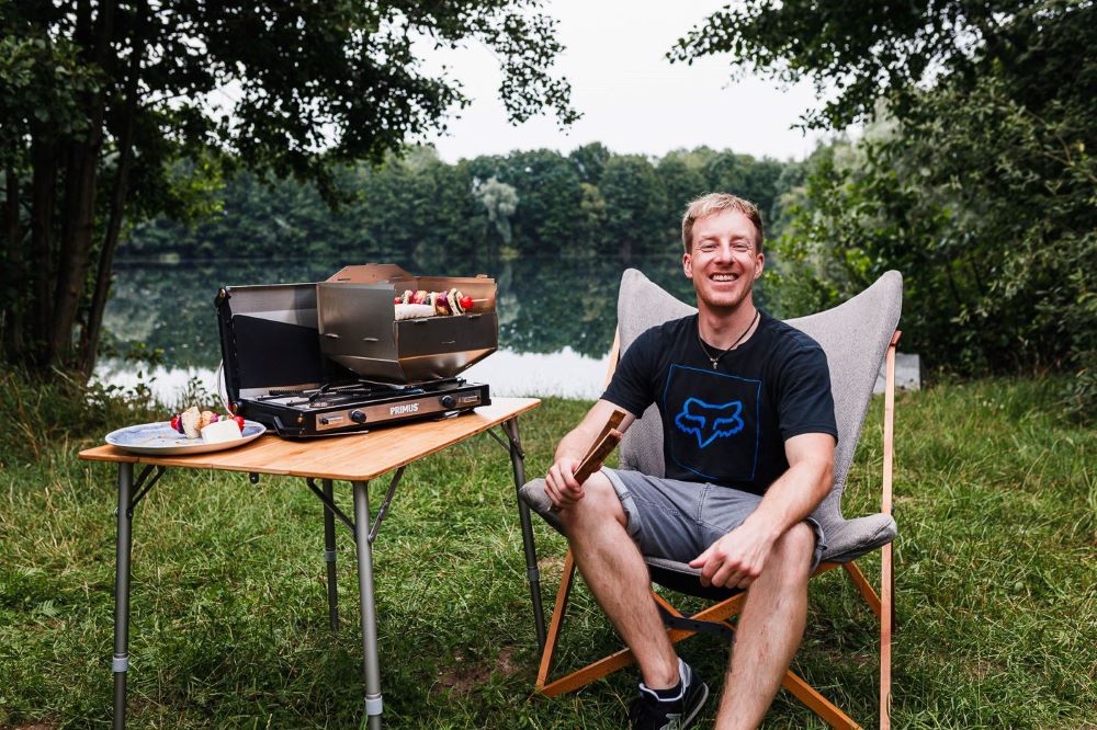 Ambientefoto:
Horst der multifunktionale Campingofen   als Gasgrill mit Gründer Fabian an einem See auf einer Wiese
